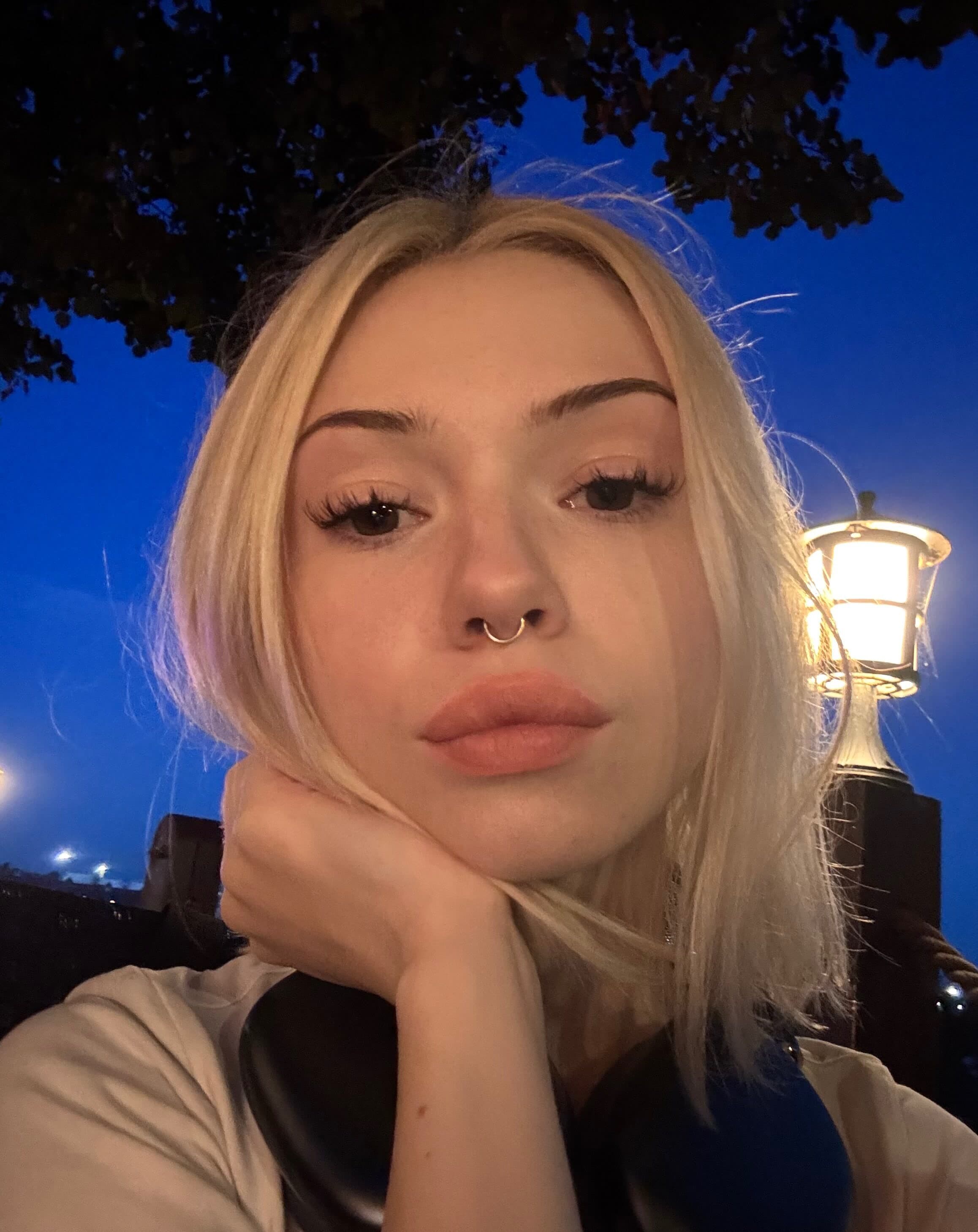 Close-up nighttime selfie of a platinum blonde woman with long straight hair, dark arched brows, dramatic winged eyeliner, thick lashes, and a gold septum ring. She rests her chin on her hand with a pout, lips slightly parted, wearing a whi