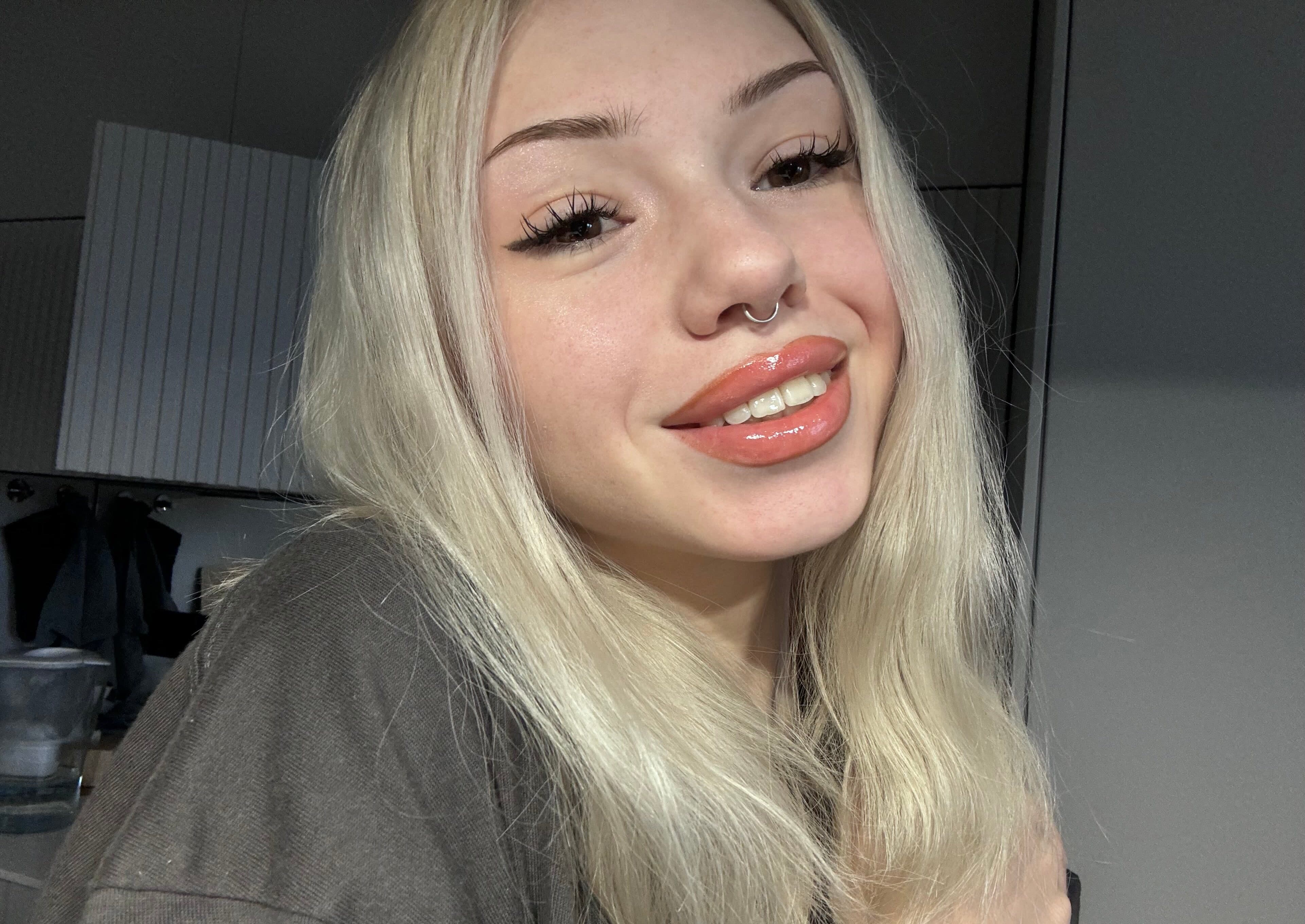 Close-up selfie of a young woman with long platinum-blonde hair falling past her shoulders, filling most of the frame. She has a septum nose ring, full glossy peach-colored lips slightly parted showing teeth in a teasing half-smile, and dra