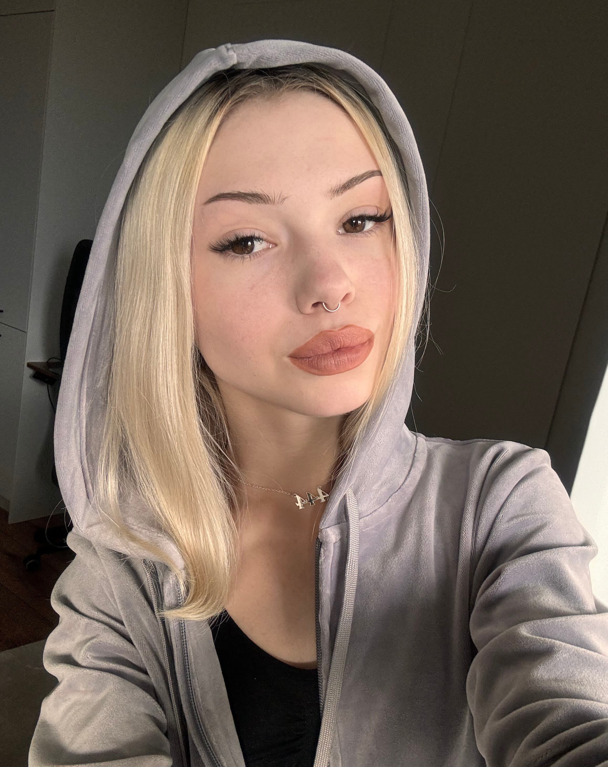 Close-up selfie of a blonde woman with the hood of a soft grey zip-up hoodie pulled up, framing her face. Her hair falls straight past her shoulders, catching warm side-light from the right. She wears a delicate gold name necklace and a bla