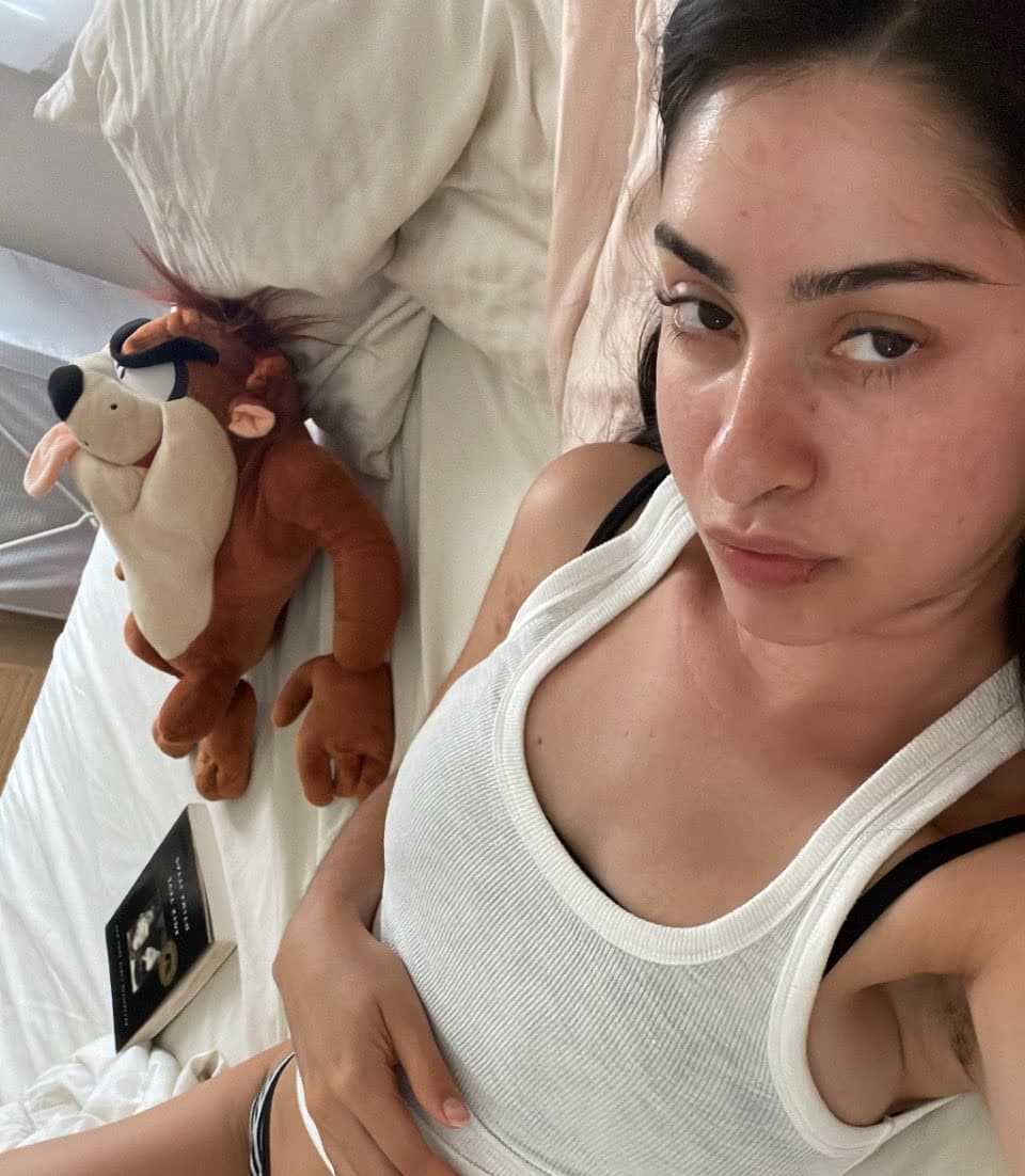 Close-up selfie of woman in white ribbed tank top with black bra straps visible, lying on bed with white linens, hand resting on hip/lower abdomen, direct eye contact with camera, intimate bedroom framing. Soft natural lighting, casual unpo