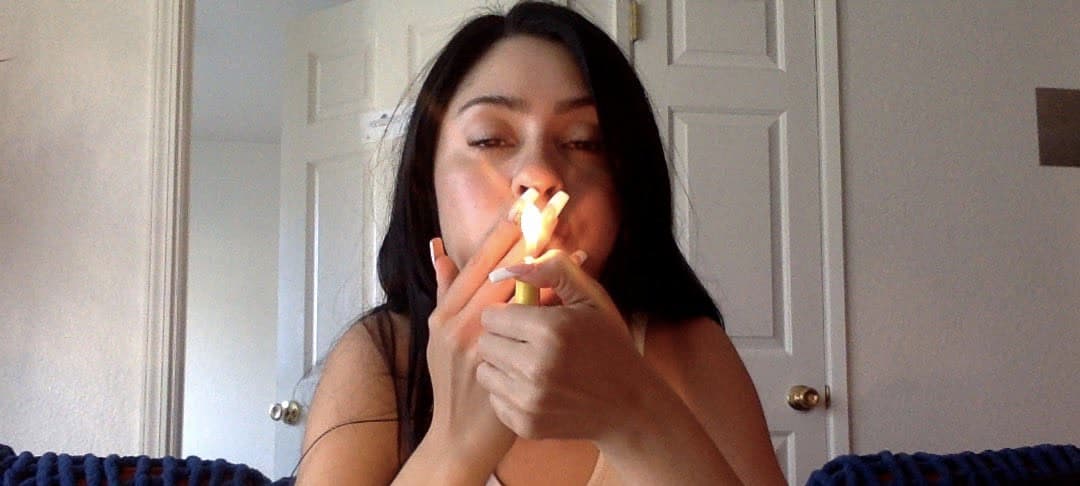 Close-up selfie of woman with long dark hair lighting a cigarette, flame illuminating her face in warm orange glow, wearing thin black spaghetti-strap top, direct eye contact with camera through half-lidded eyes, intimate framing from chest