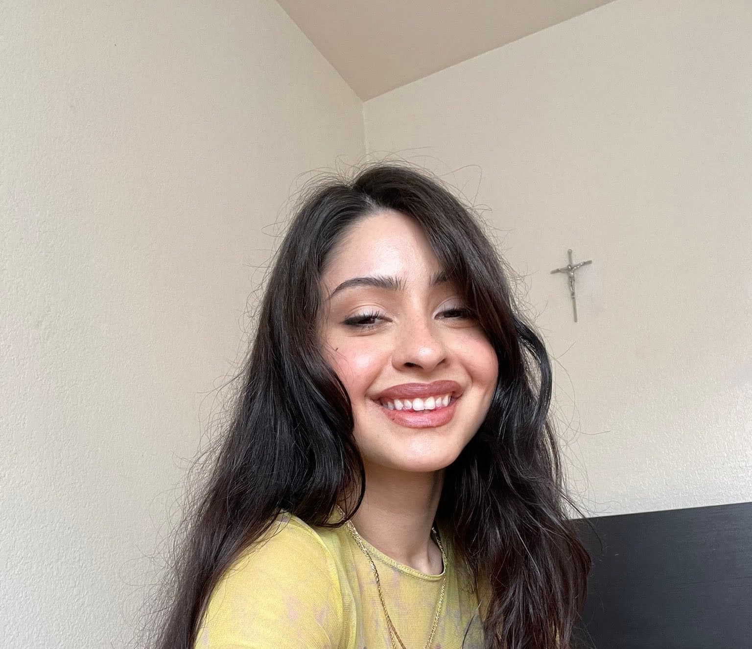 Close-up selfie of a young woman with long dark wavy hair cascading past shoulders, wearing a soft yellow semi-sheer top and layered gold necklaces. Head tilted slightly, warm smile with teeth visible, eyes relaxed and inviting. Shot from s