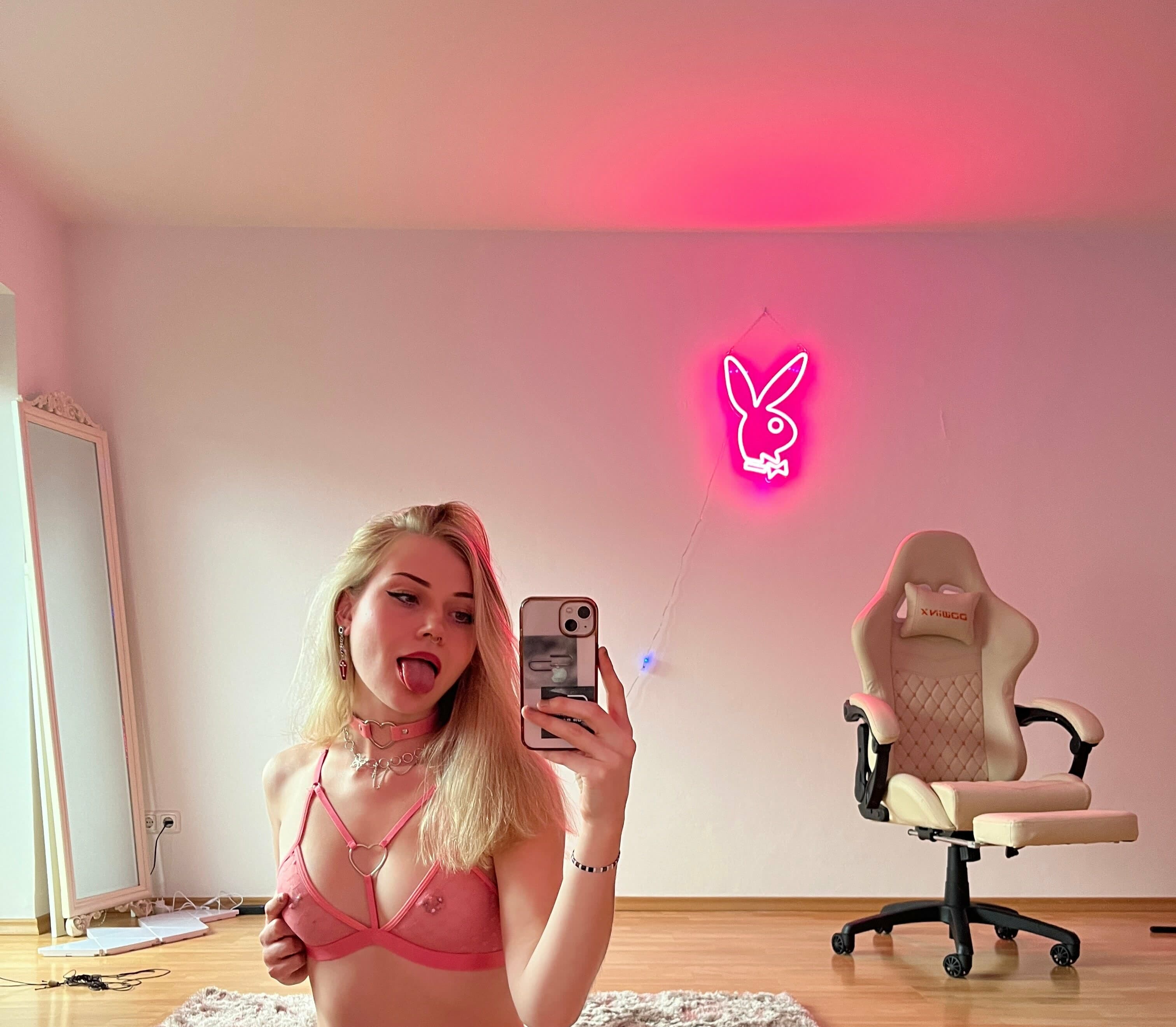 Blonde woman taking mirror selfie, tongue playfully extended toward camera with direct eye contact. Wearing pink strappy lingerie bra with heart-shaped metal hardware at center chest, pink choker with heart ring detail, layered chain neckla