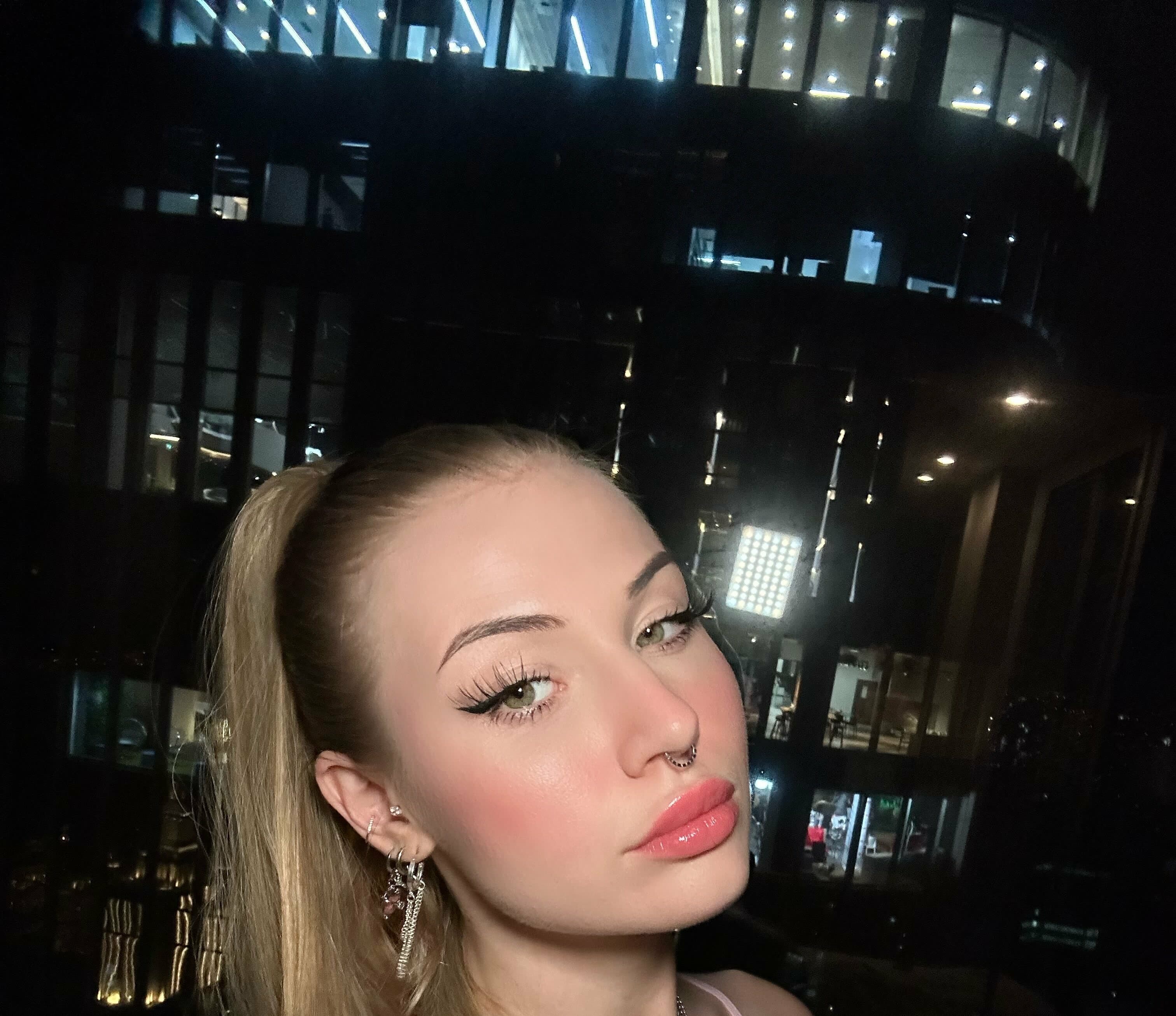 Close-up portrait of a woman with blonde hair pulled into a high ponytail, wearing dangling chain earrings and a nose ring. Face turned slightly to the side with direct gaze toward camera, glossy pink lips, dramatic winged eyeliner, flushed
