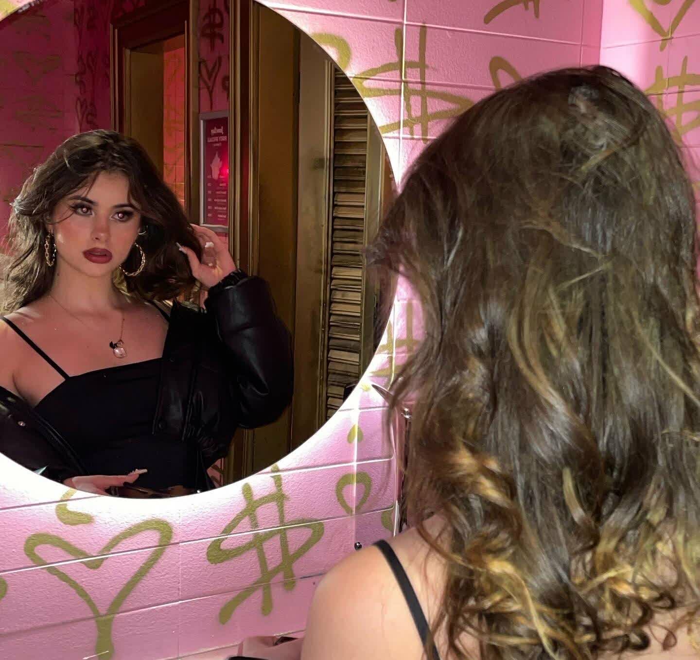 Mirror selfie of a woman with voluminous dark wavy hair with caramel highlights, seen from behind and in reflection. She wears a black spaghetti-strap top with a black leather jacket slipping off one shoulder, revealing decolletage. Large g