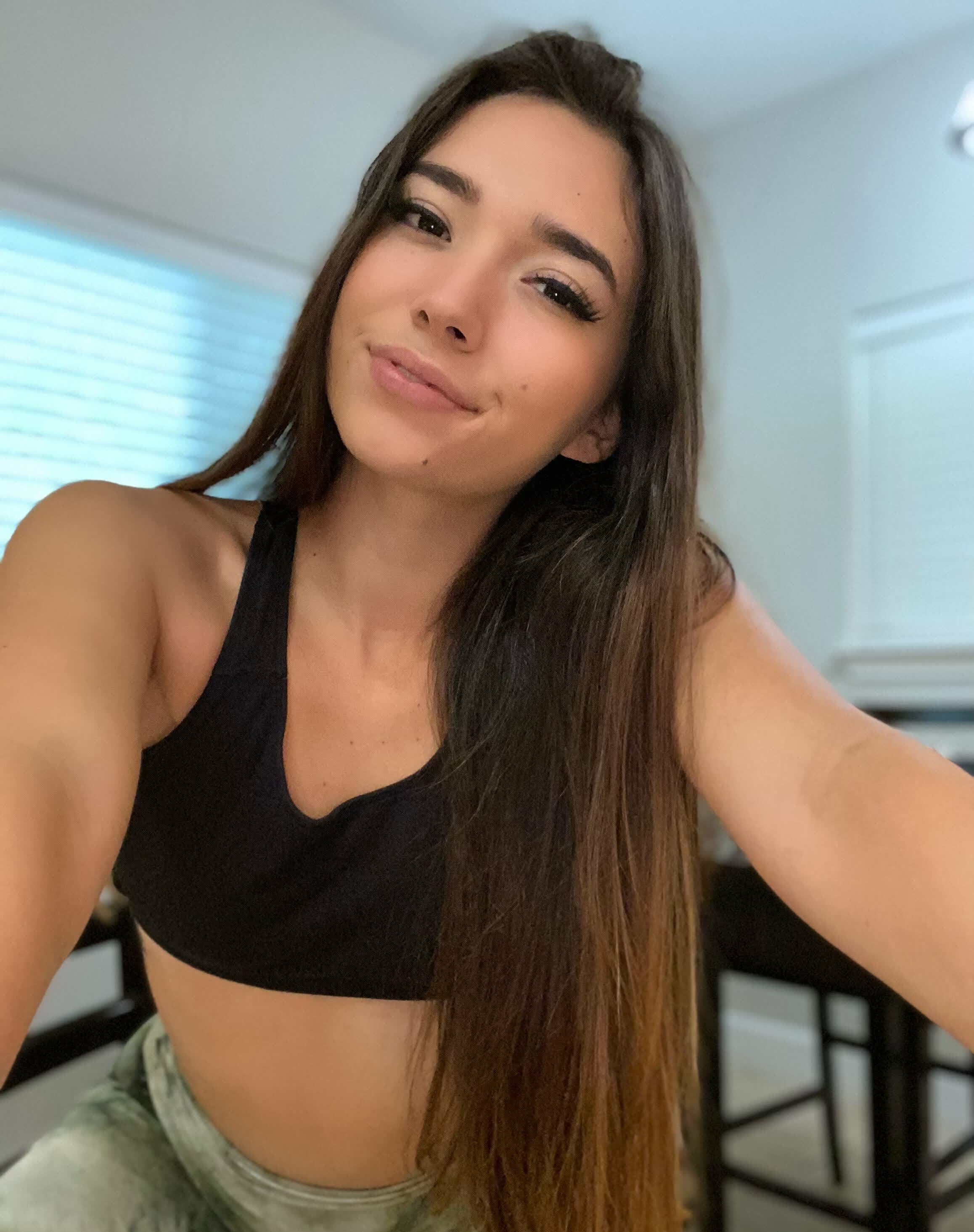 Selfie shot of a young woman with long brown-to-blonde ombre hair, wearing a black sports bra with deep V-neckline and light green/grey high-waisted bottoms. Arm extended holding camera, slight smile with full lips, defined brows, long lash