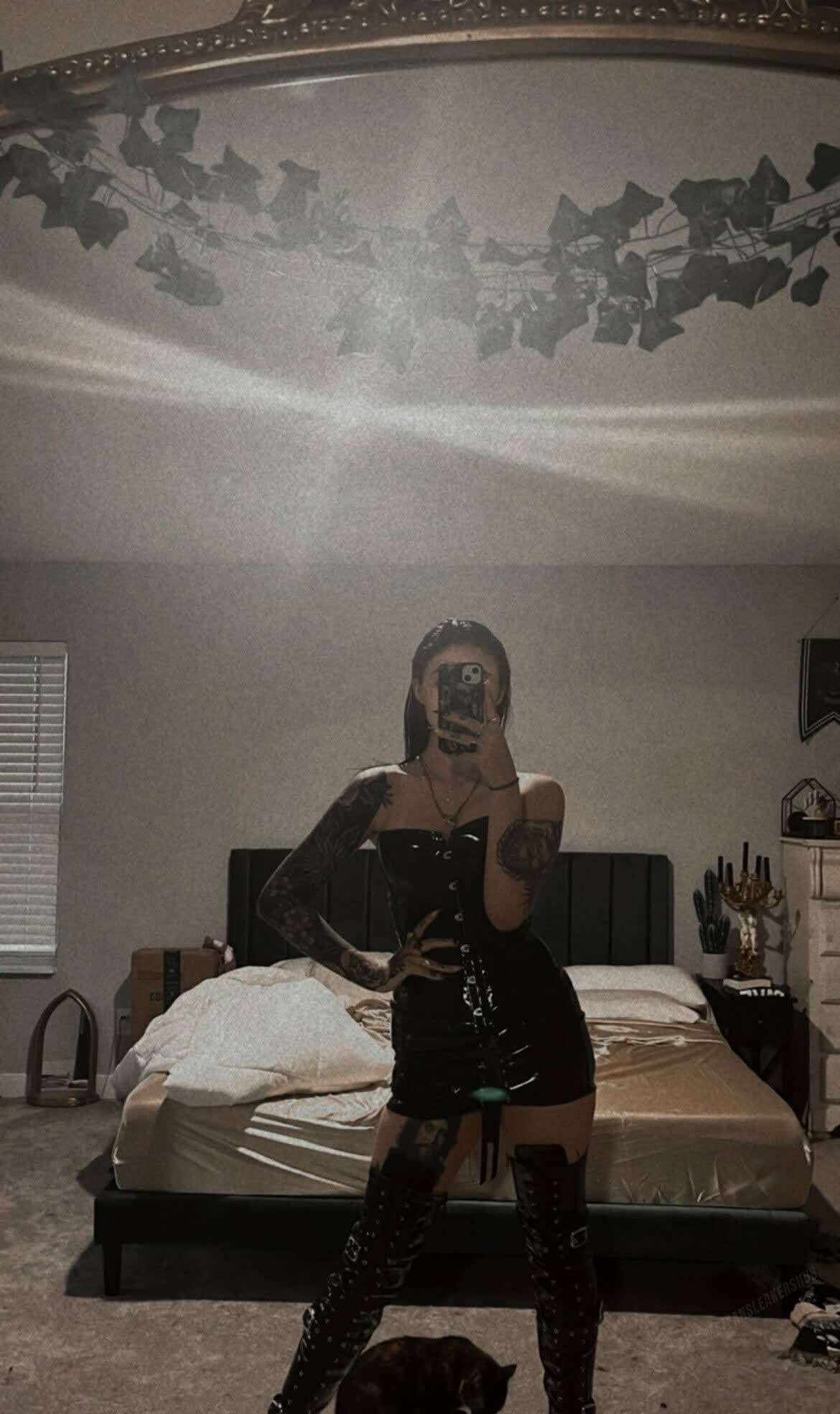 Full-length mirror selfie of a tattooed woman in a dim bedroom, standing with legs apart and one hand on hip, the other holding phone to obscure her face. She wears a strapless black PVC mini dress with front lacing/buckles and thigh-high b