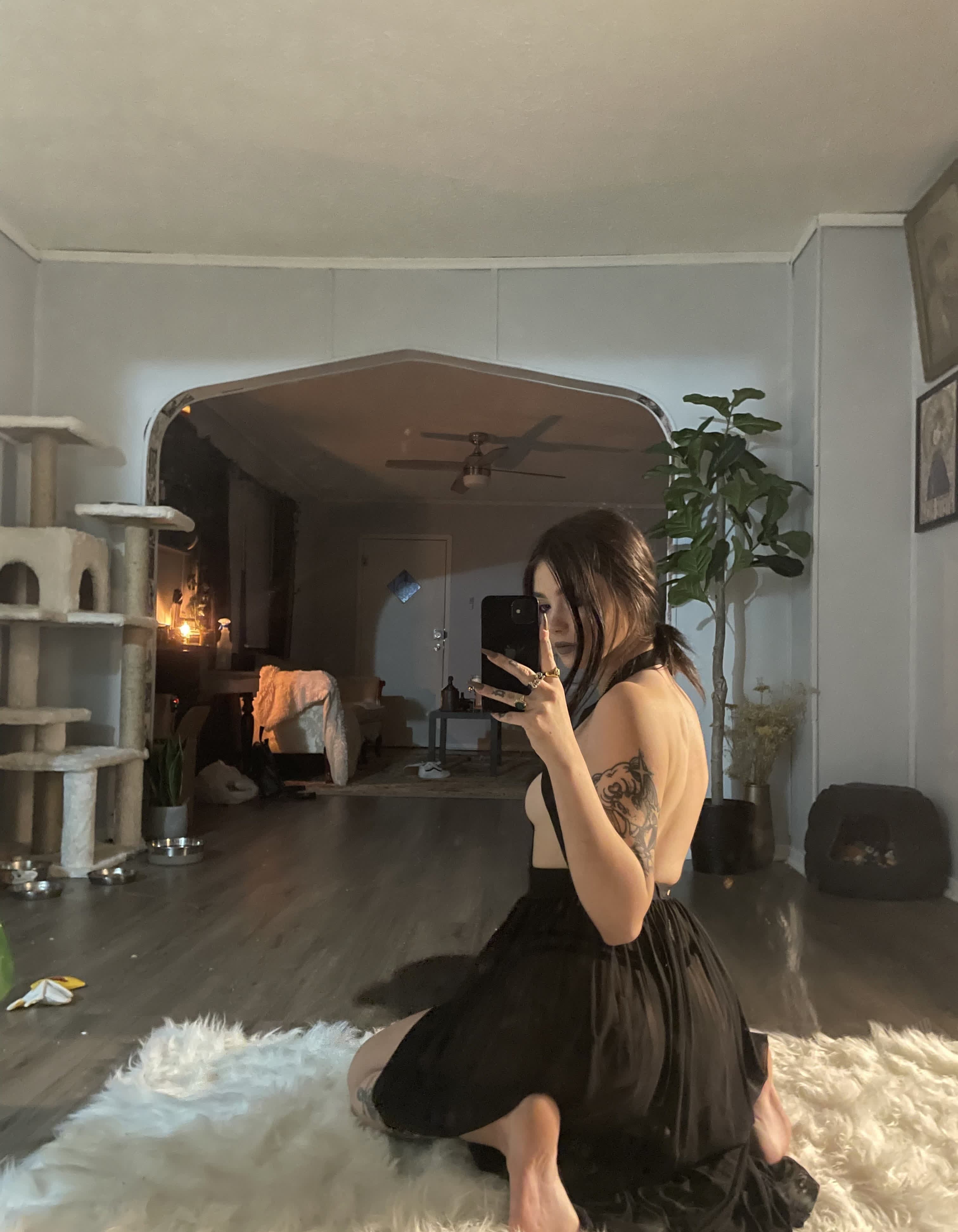 Mirror selfie of a woman kneeling on a white fluffy rug, shot from behind at a three-quarter angle. She wears a black backless dress with thin straps, sideboob visible, the fabric pooling around her knees. Dark hair in a loose ponytail, lon