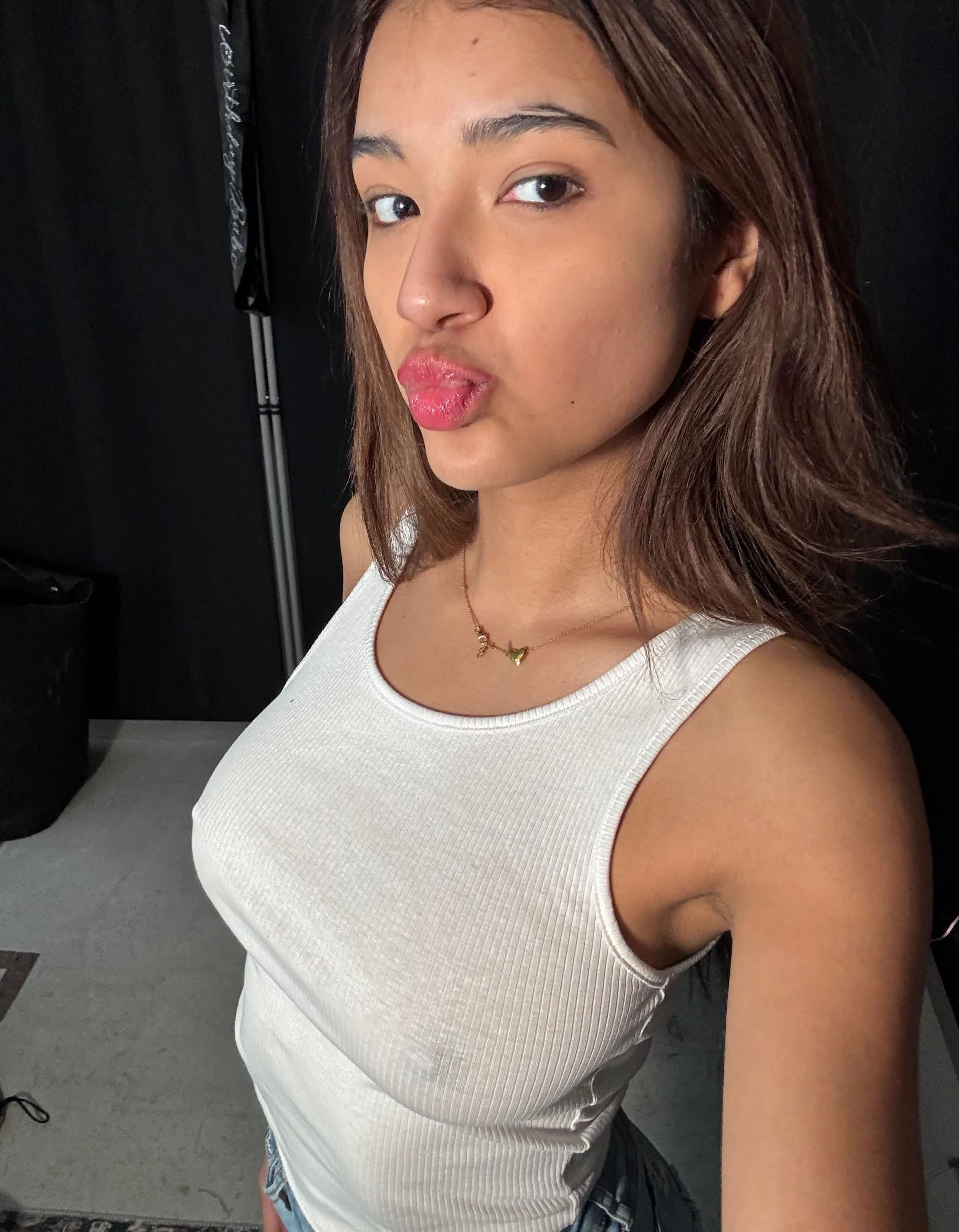 Close-up selfie of a young woman with shoulder-length brown hair, wearing a tight white ribbed tank top that hugs her chest, and a delicate gold butterfly necklace. She is puckering her lips in a kiss pose, looking directly at the camera wi