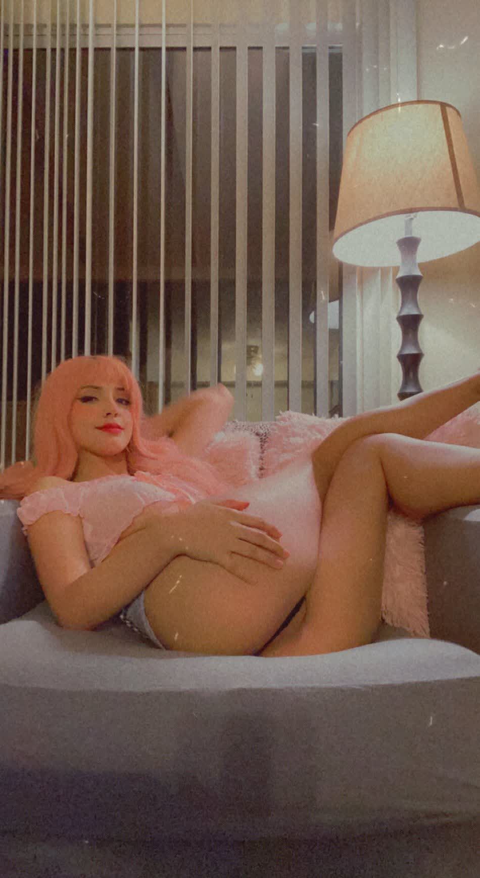 Pink-haired figure reclining on a light gray sofa, wearing a soft pink ruffled off-shoulder top and light blue/gray underwear. One arm rests behind the head, the other hand placed low on the inner thigh, drawing attention to the hip and leg