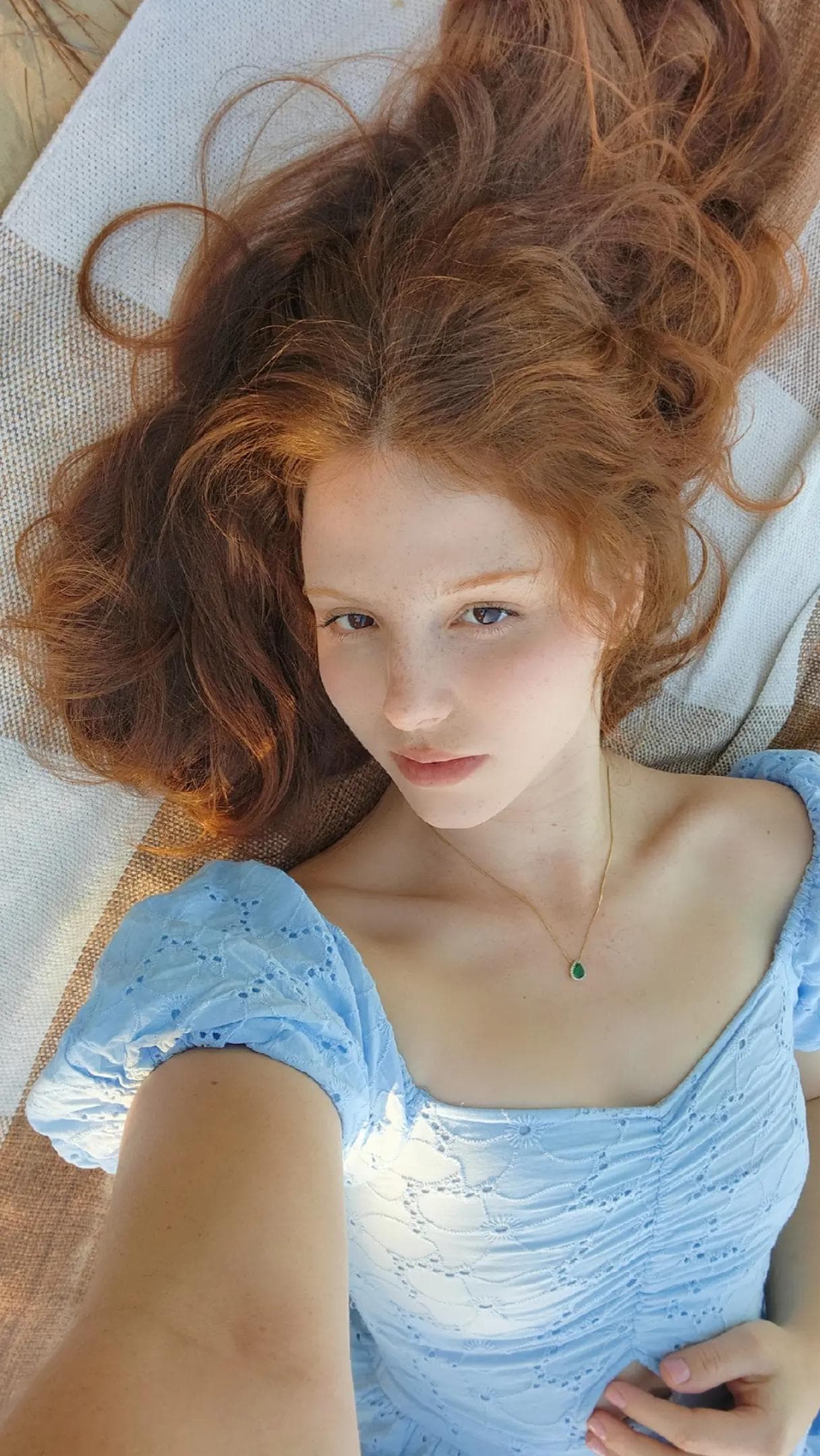 Overhead selfie of a young woman with voluminous wavy red/auburn hair spread across a light fabric surface. She wears a light blue eyelet-lace top with puff sleeves and square neckline that exposes collarbones and upper chest. A delicate go