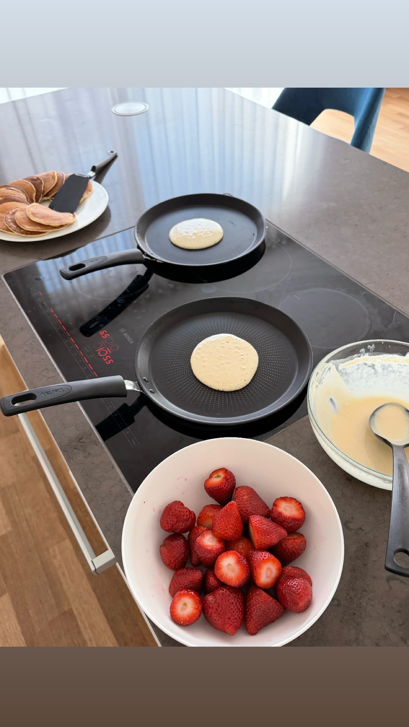 Overhead kitchen scene showing pancake preparation on an induction cooktop. Two black frying pans with pancakes at different cooking stages, a bowl of fresh halved strawberries in the foreground, a glass bowl with pancake batter and ladle t