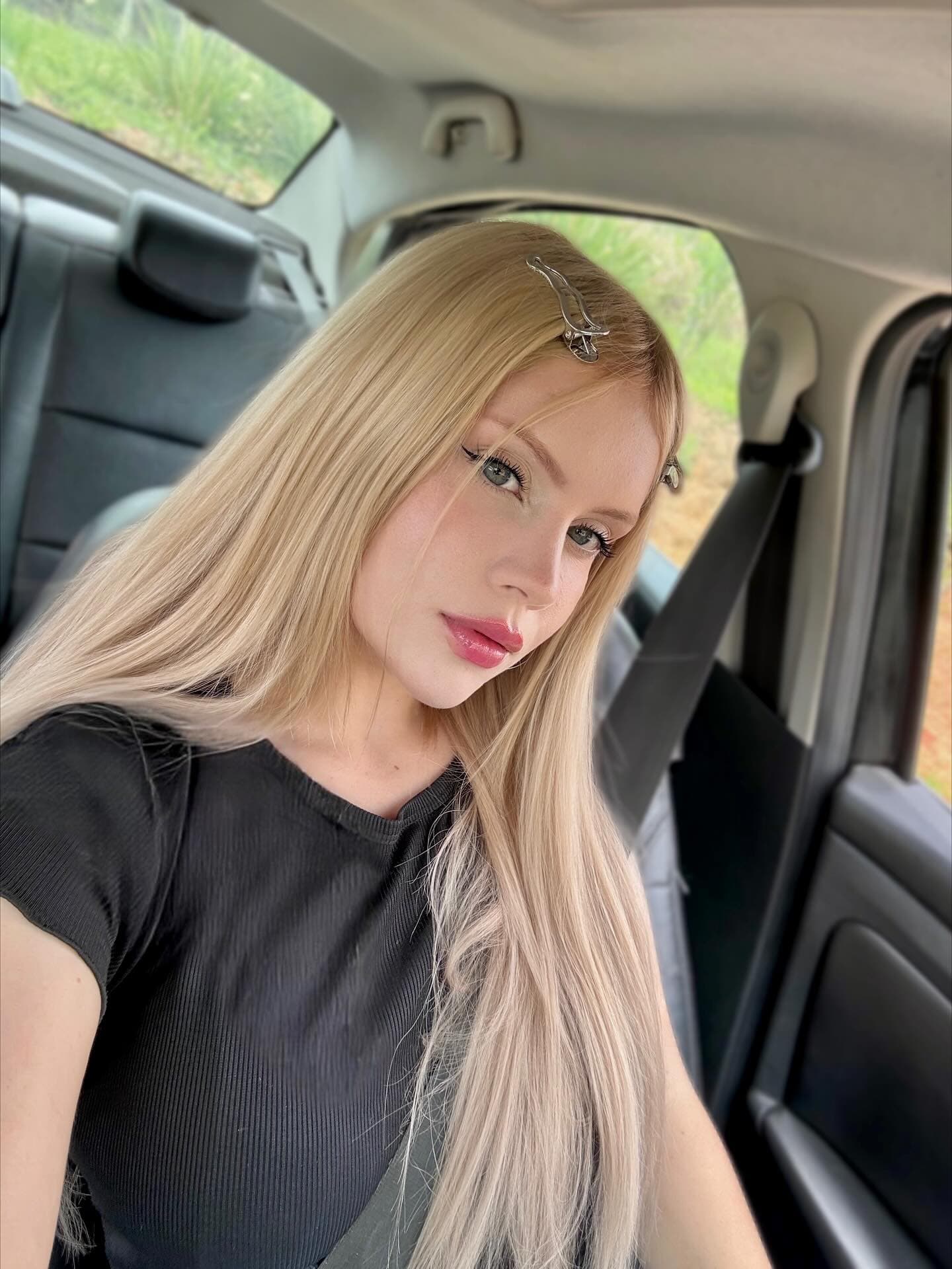 Close-up car selfie of a young woman with very long platinum blonde hair, two decorative silver hair clips visible on one side. She wears a fitted black short-sleeve ribbed top and a seatbelt crossing her chest. Shot from slightly above, he