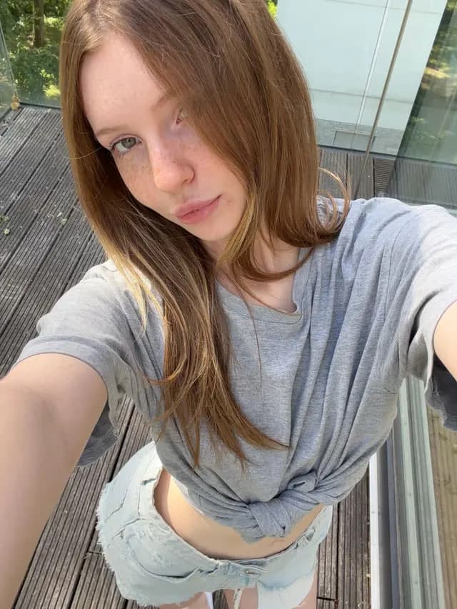 Selfie of a young woman with long light-brown hair and blue eyes, freckles visible across her nose and cheeks. She wears a heather grey t-shirt knotted at the waist to expose her midriff, paired with light-wash denim cutoff shorts. Shot fro