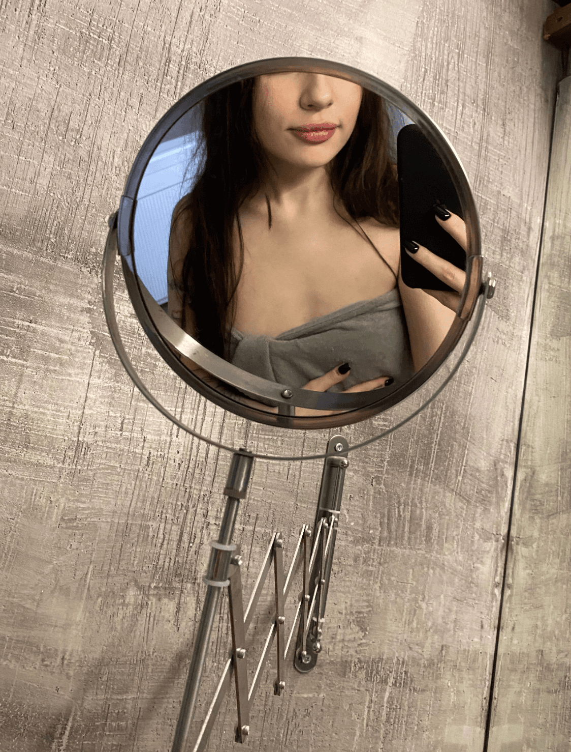 Mirror selfie of a young woman with long dark hair, face partially cropped at forehead, visible from nose down. Wrapped in a grey towel or strapless top, one hand holding a black phone with black nail polish, the other hand resting at her w