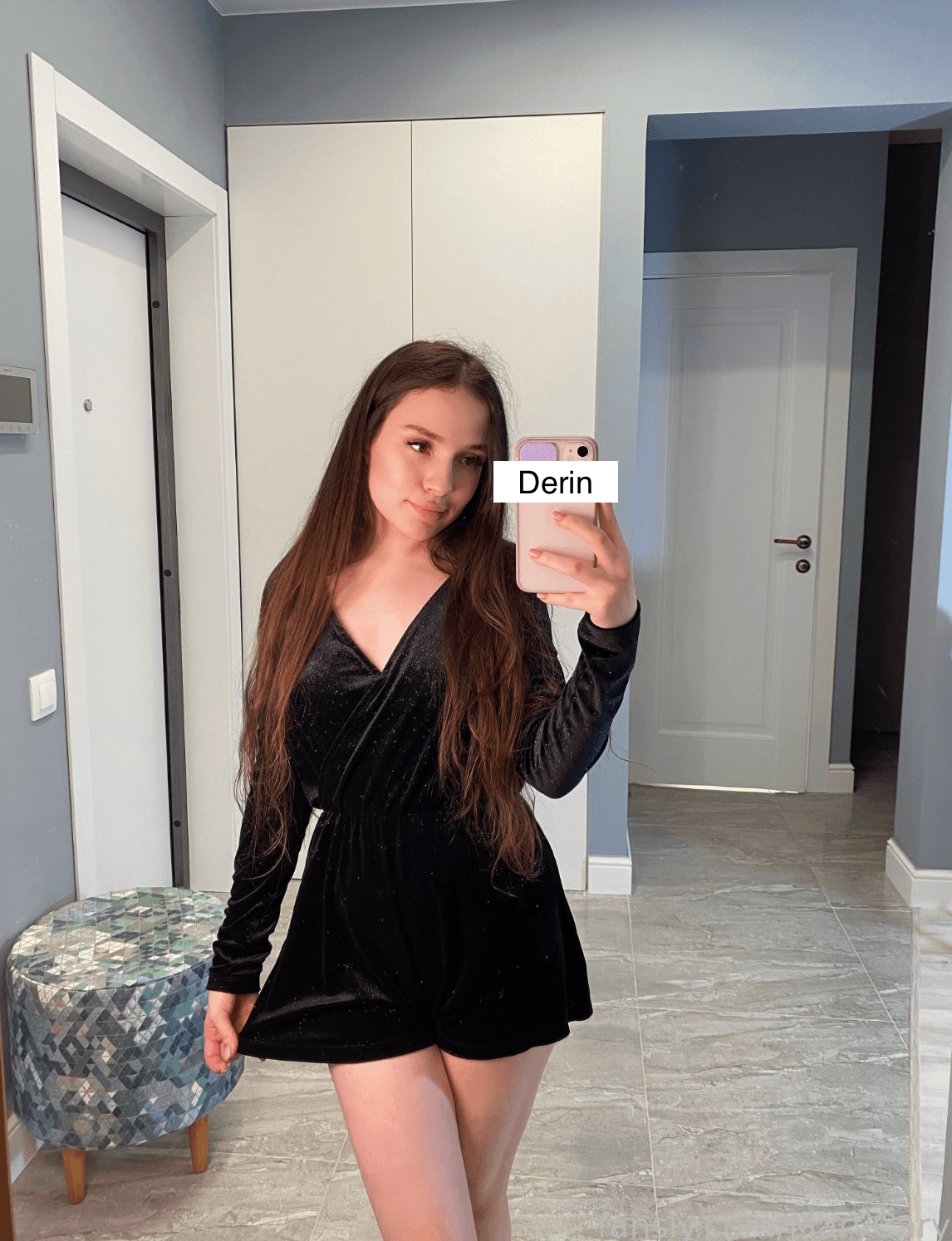 Mirror selfie of a young woman with very long straight brown hair, wearing a short black velvet wrap dress with deep V-neckline and long sleeves. She holds a rose-gold phone at chest level, slight hip cock, legs crossed at ankle, neutral to