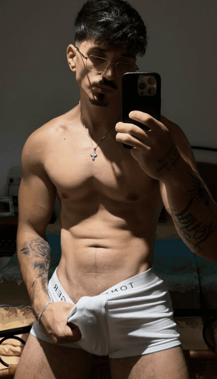 Shirtless muscular man with defined abs and chest taking a mirror selfie in dim lighting. Wearing white boxer briefs with visible waistband lettering, pulling the front down slightly with one hand to reveal lower abs and hint at more. Gold
